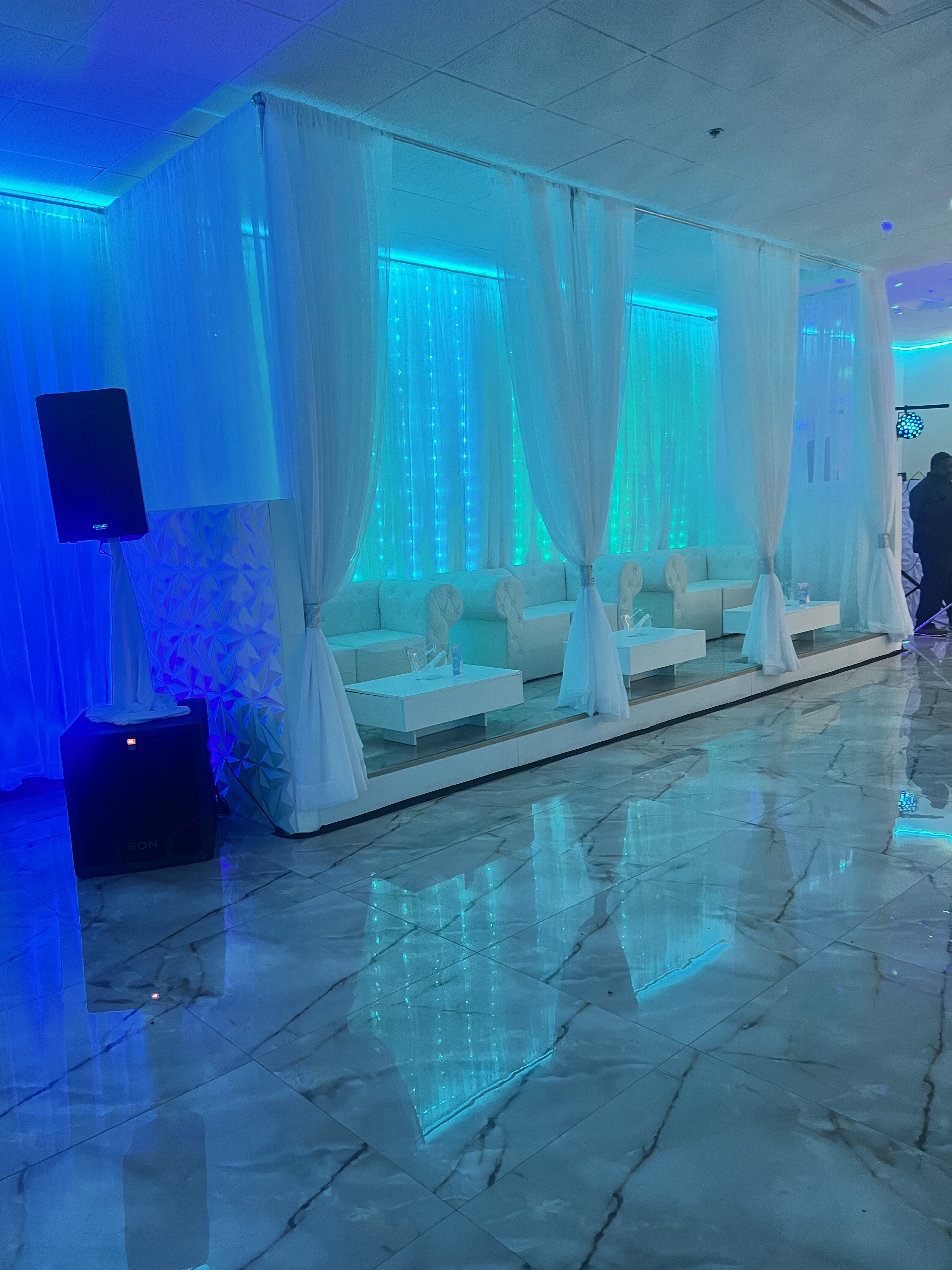 A modern event space with blue lighting, white drapery, and glossy marble floors reflecting the blue lights. white benches with head sculptures line the wall.
