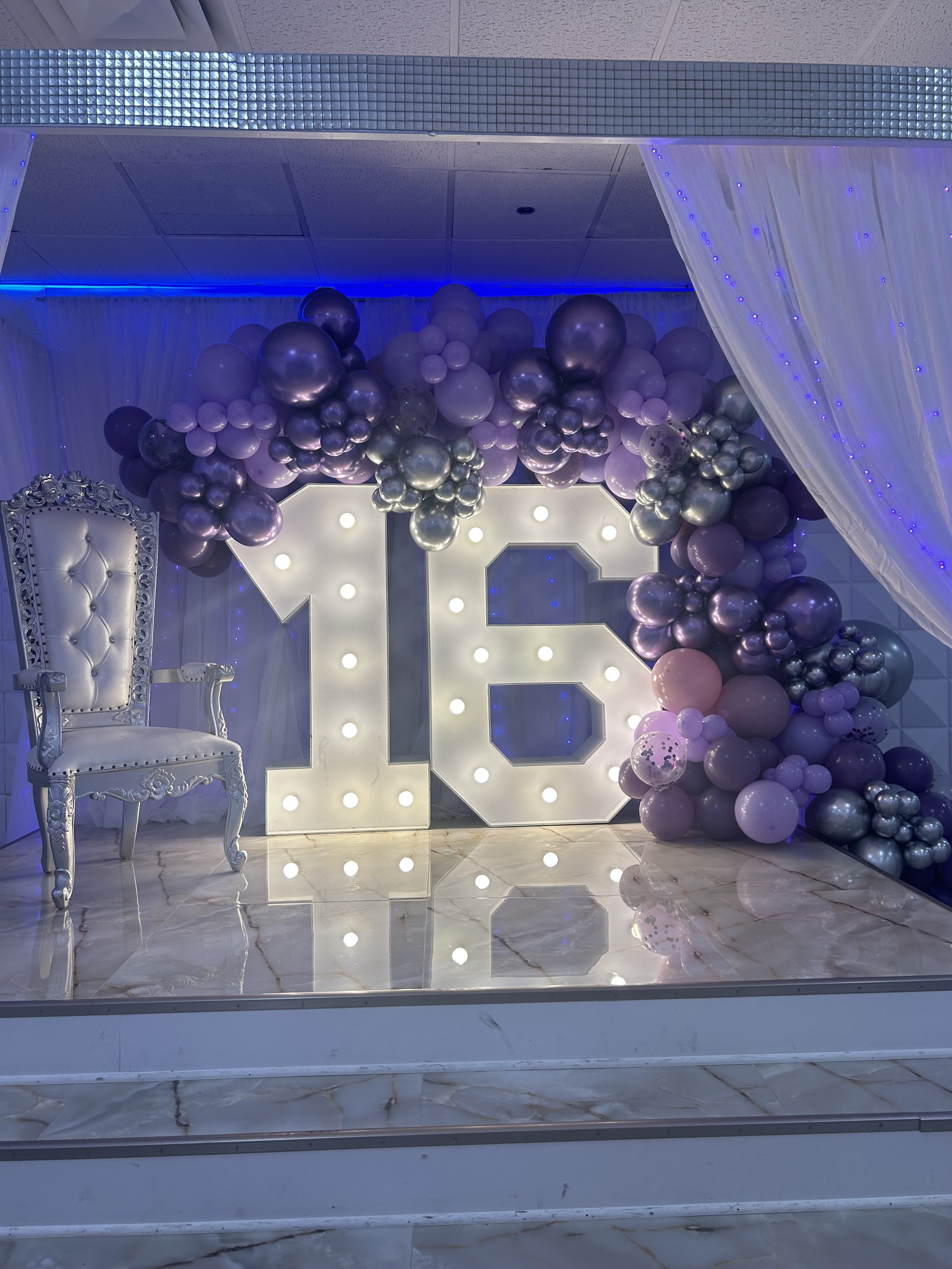 A festive setup for a quinceañera featuring a large "15" light-up sign, surrounded by clusters of purple and grey balloons, with a white ornate chair on the left, all highlighted by soft blue lighting.