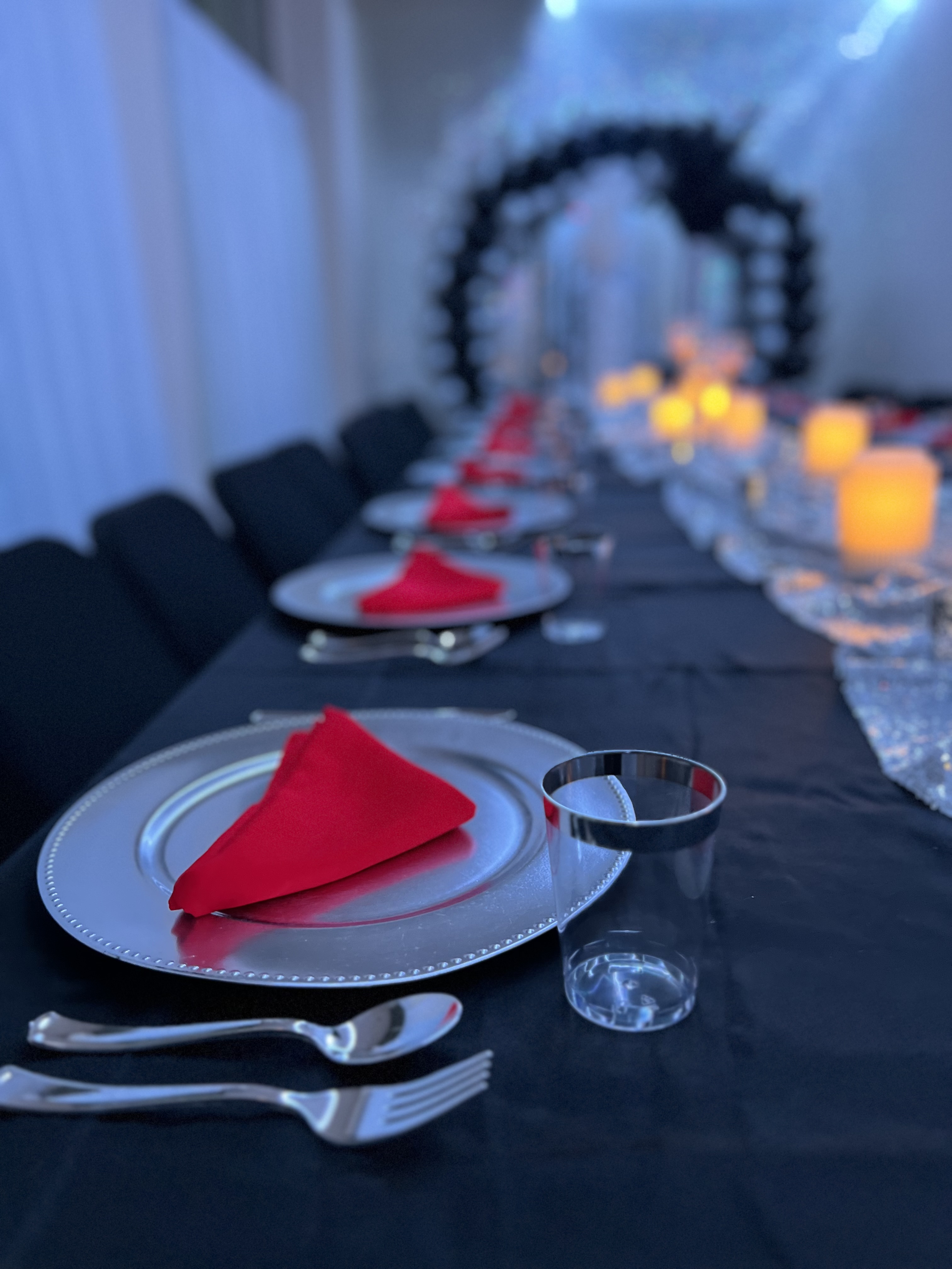 An elegantly set dinner table with a focus on a red napkin on a silver plate, complemented by silverware, glasses, and candle lights, leading to a balloon arch in the background.