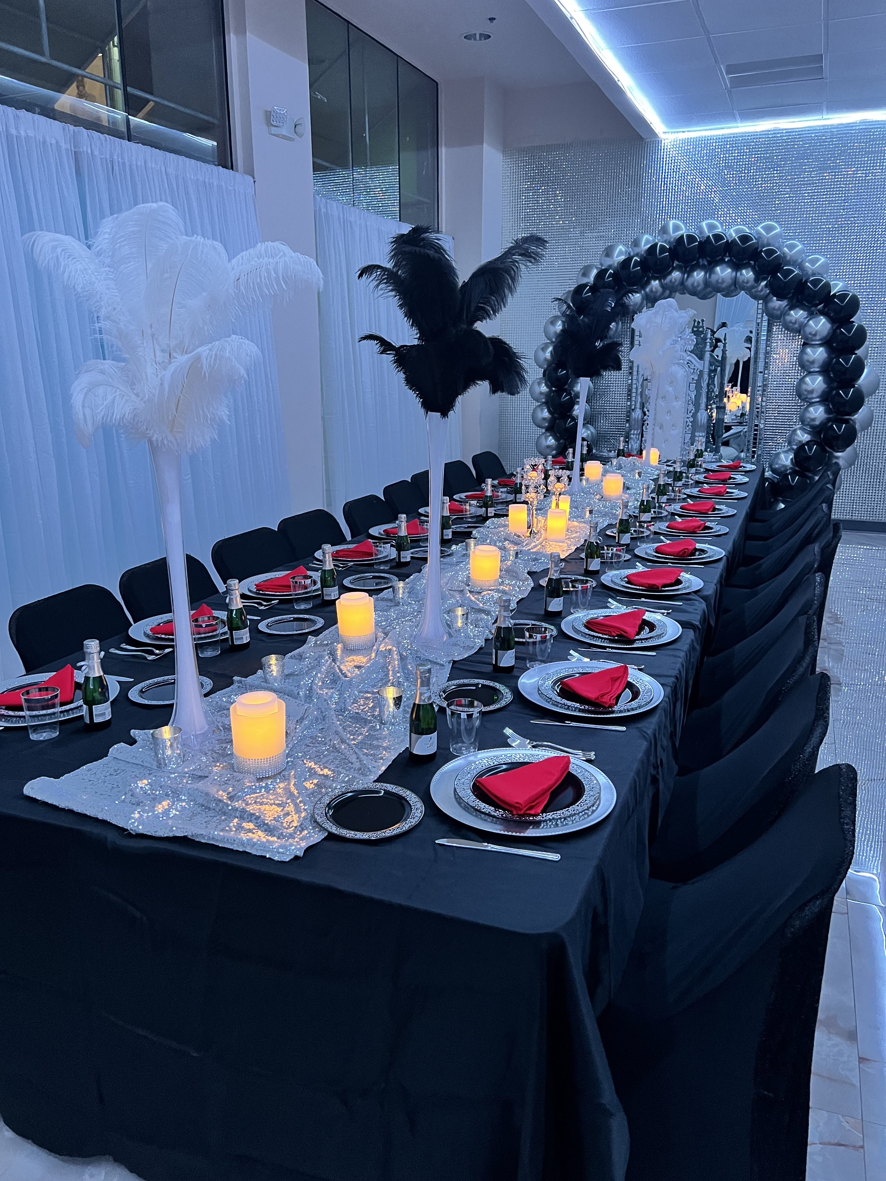 Elegant dining table setup with black tablecloth, red napkins, and white and black feather centerpieces. candles and mirrors add ambiance to the festive decor.