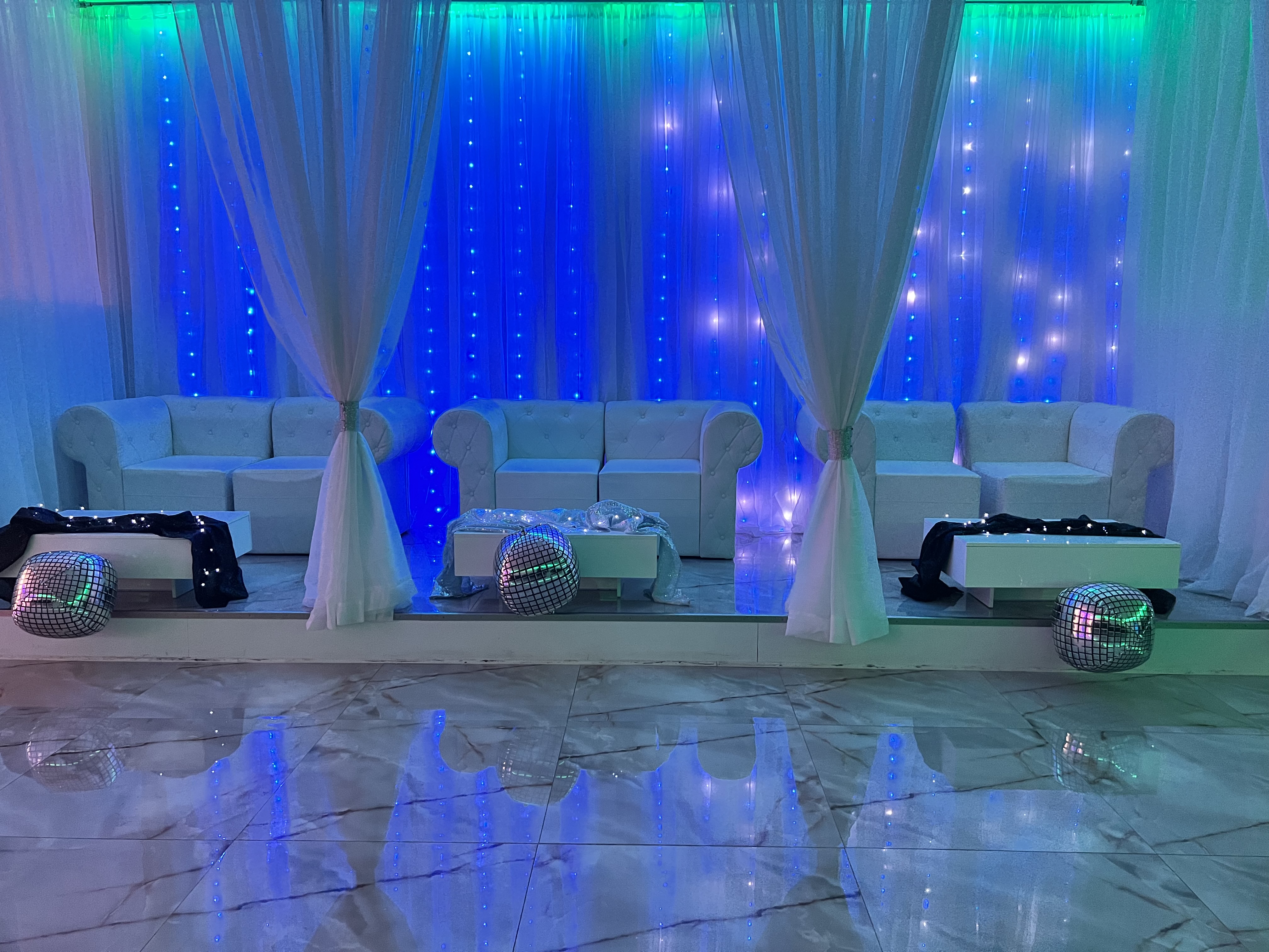 A posh event space with a blue-lit back wall, plush white sofas, shimmery curtains, and disco balls on the floor reflecting light.