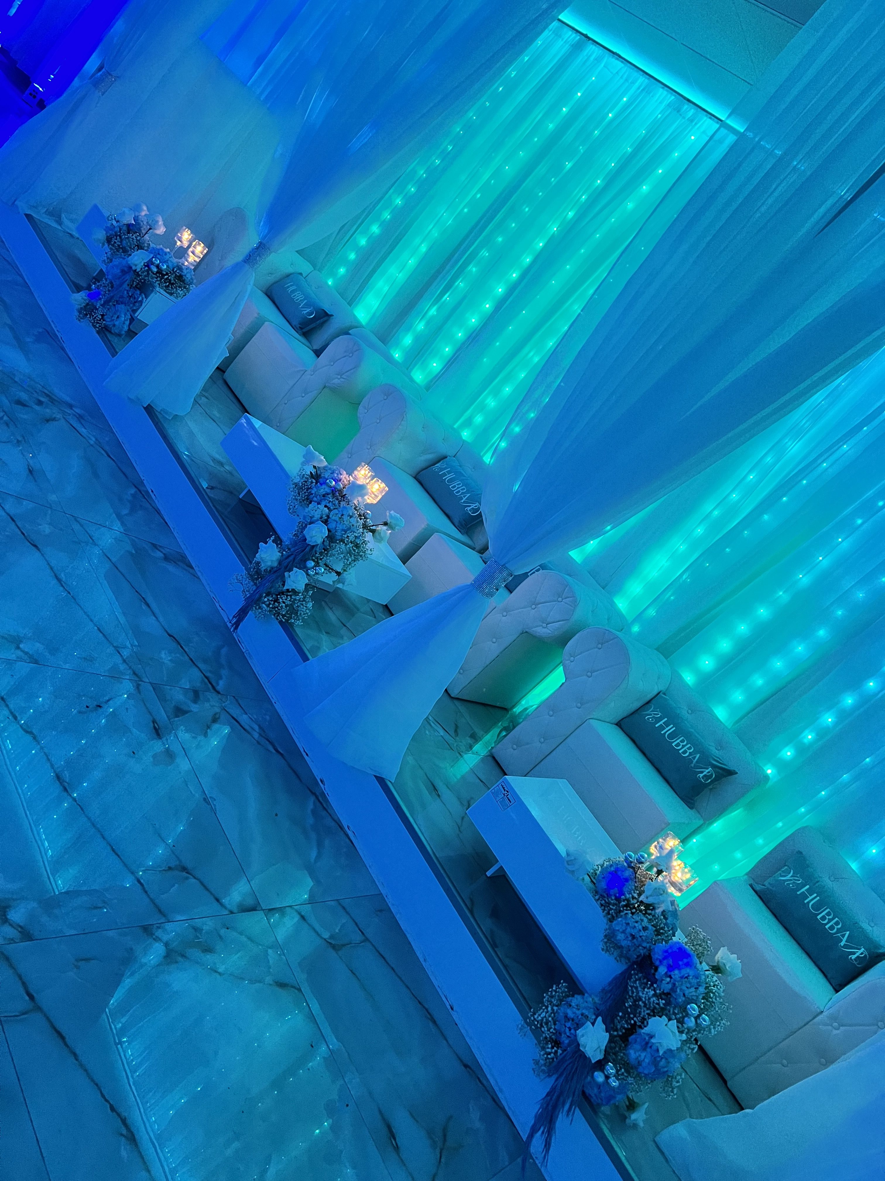An elegantly decorated event space with a blue lighting theme, featuring white sofas, blue and white floral arrangements, and illuminated vertical light panels.
