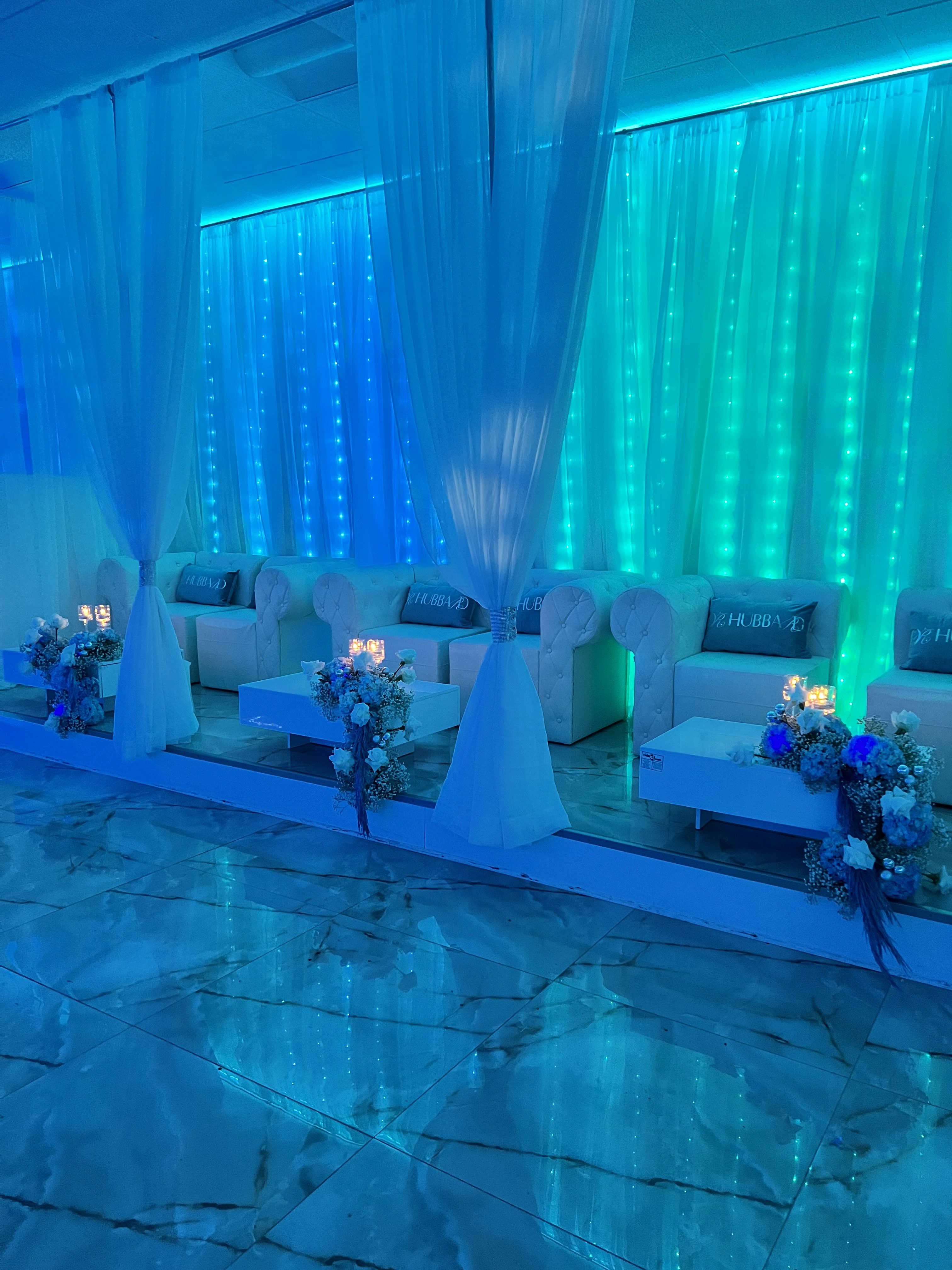An elegant event space with blue lighting, featuring white sofas adorned with floral arrangements, marble floors, and draped white curtains.