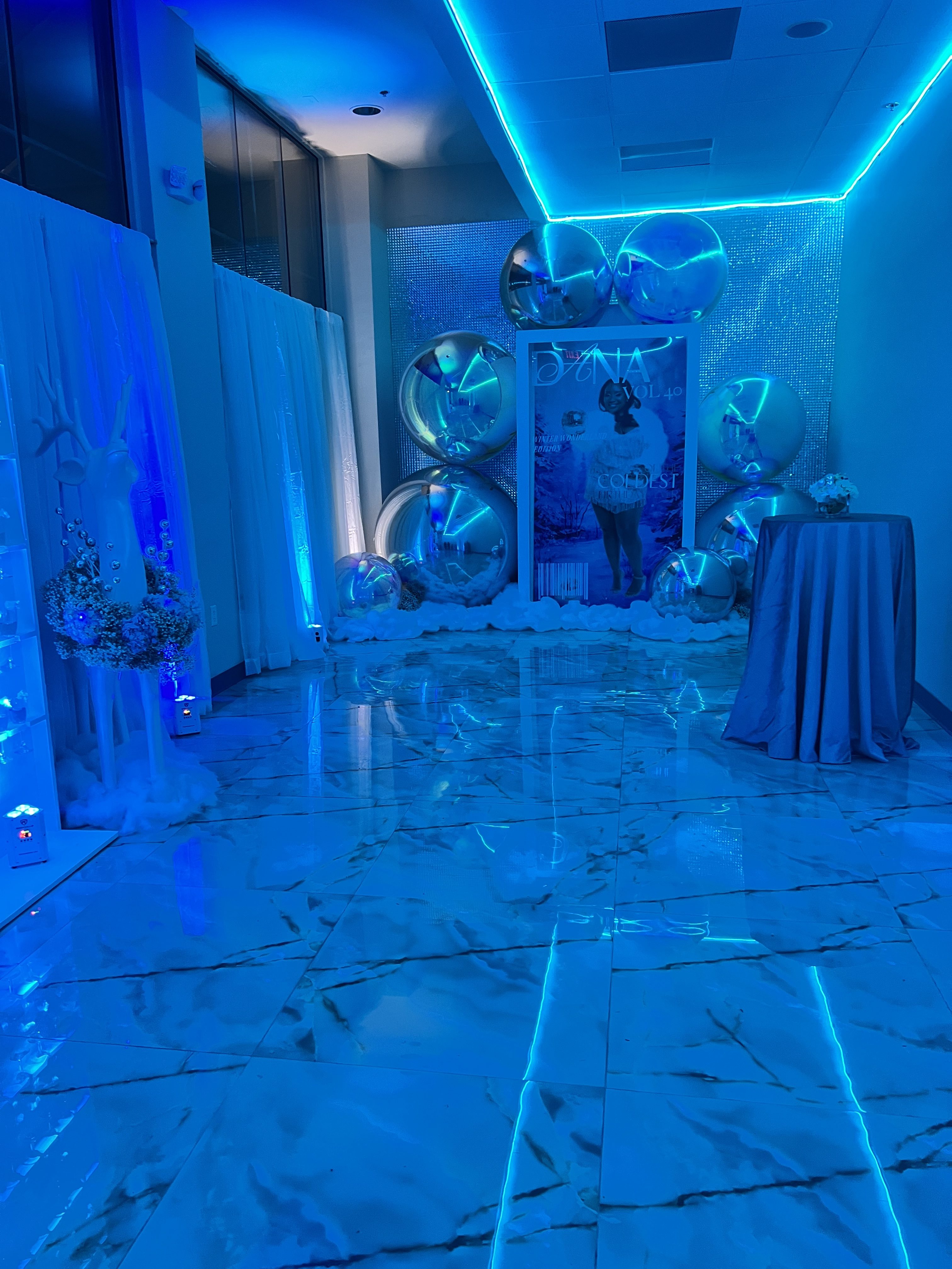 A blue-lit venue entrance with silver balloons, draped fabrics, and a poster titled "dani" flanked by statues and floral arrangements. the floor reflects the vibrant lighting setup.