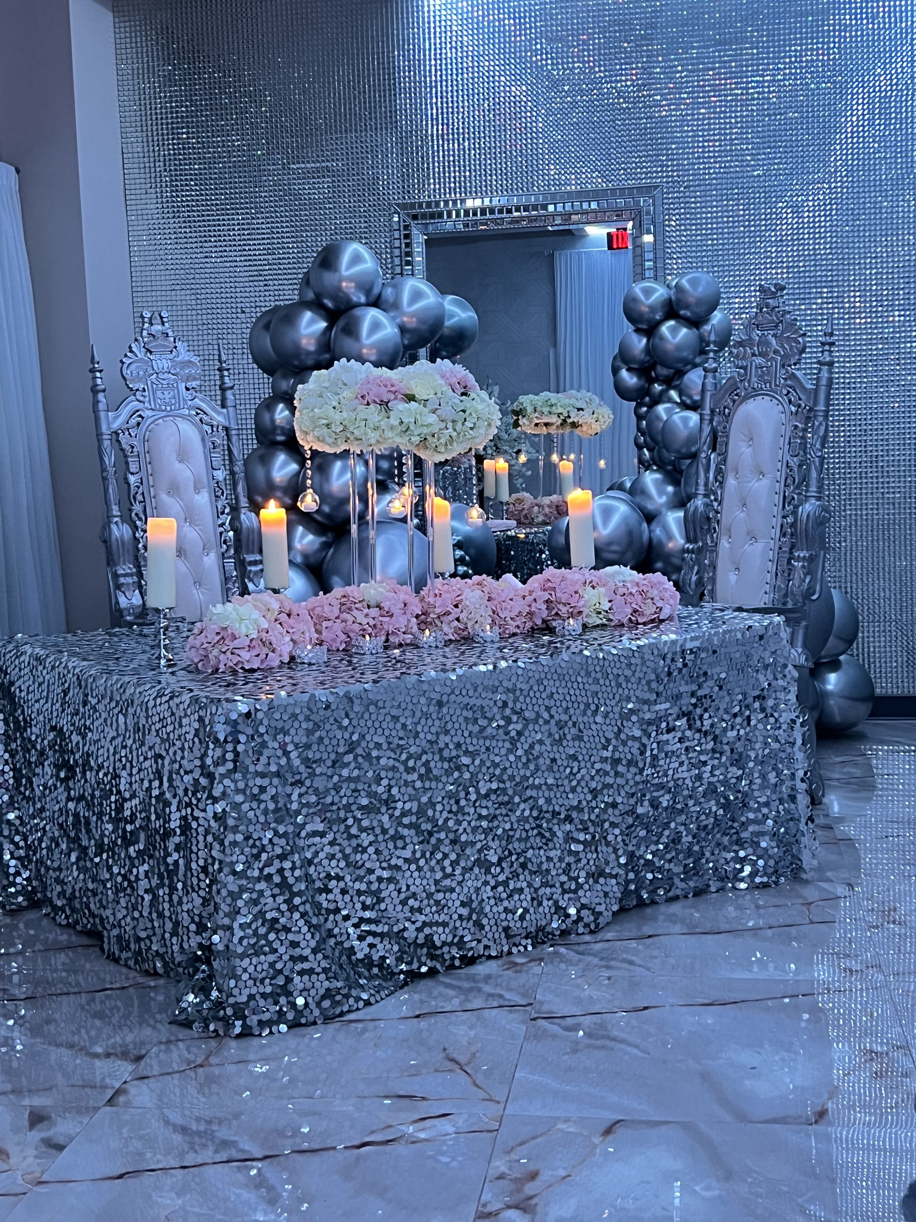 A lavish event setting with two ornate white chairs, a table covered in shimmering silver and pink flowers, surrounded by lit candles, balloons, and a sparkly backdrop.