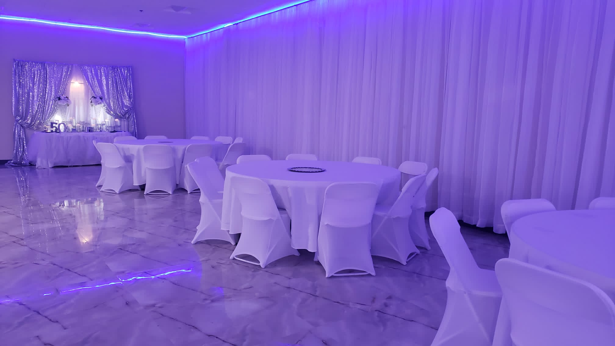 An elegantly decorated event hall bathed in purple lighting, featuring round tables with white cloth covers, and a shimmering curtain backdrop with a "50th" sign.