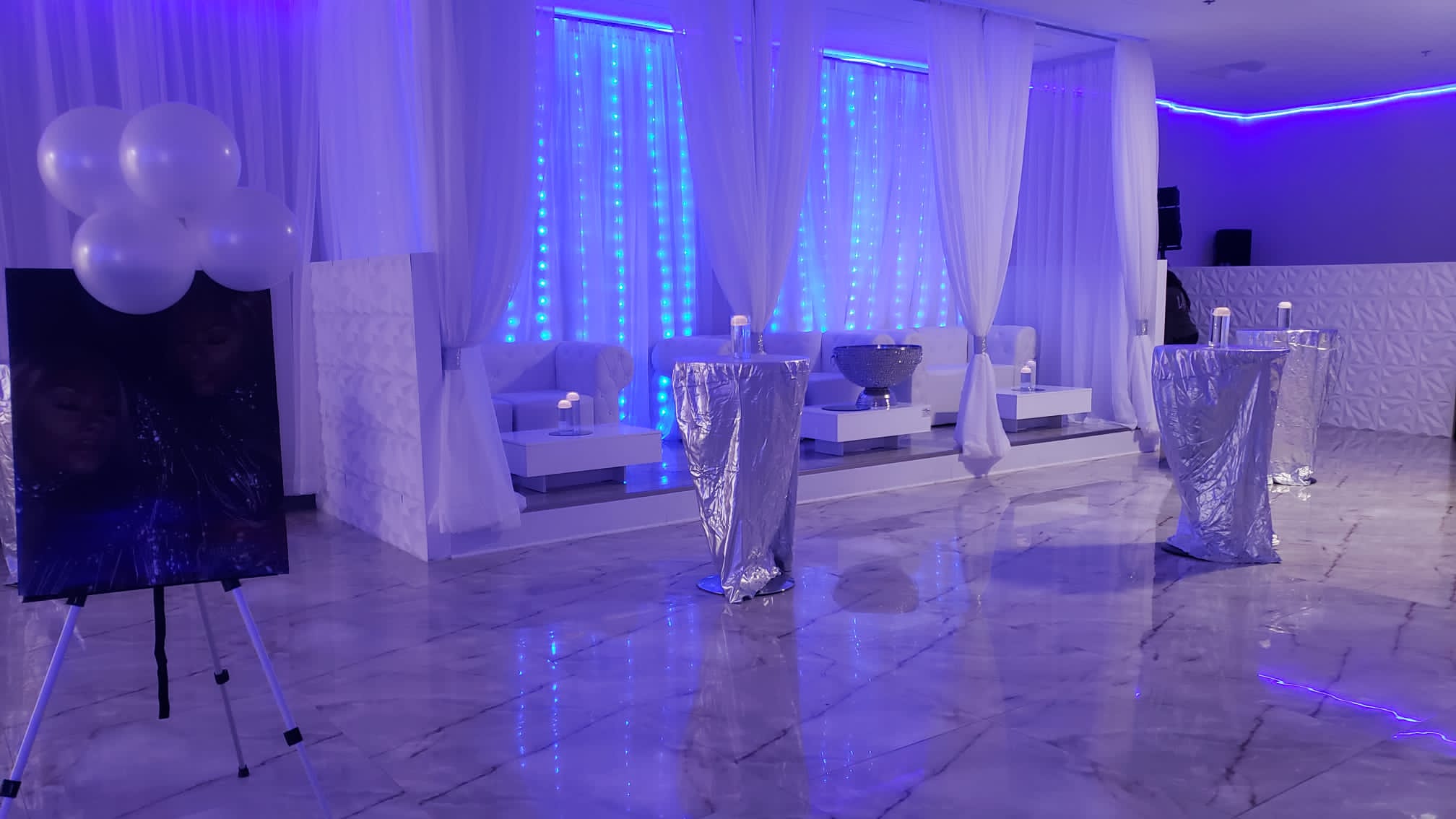 A modern event space lit with blue led lights, featuring white draped curtains, silver high-top tables, and white lounge seating. balloons and portrait canvases add to the decor.
