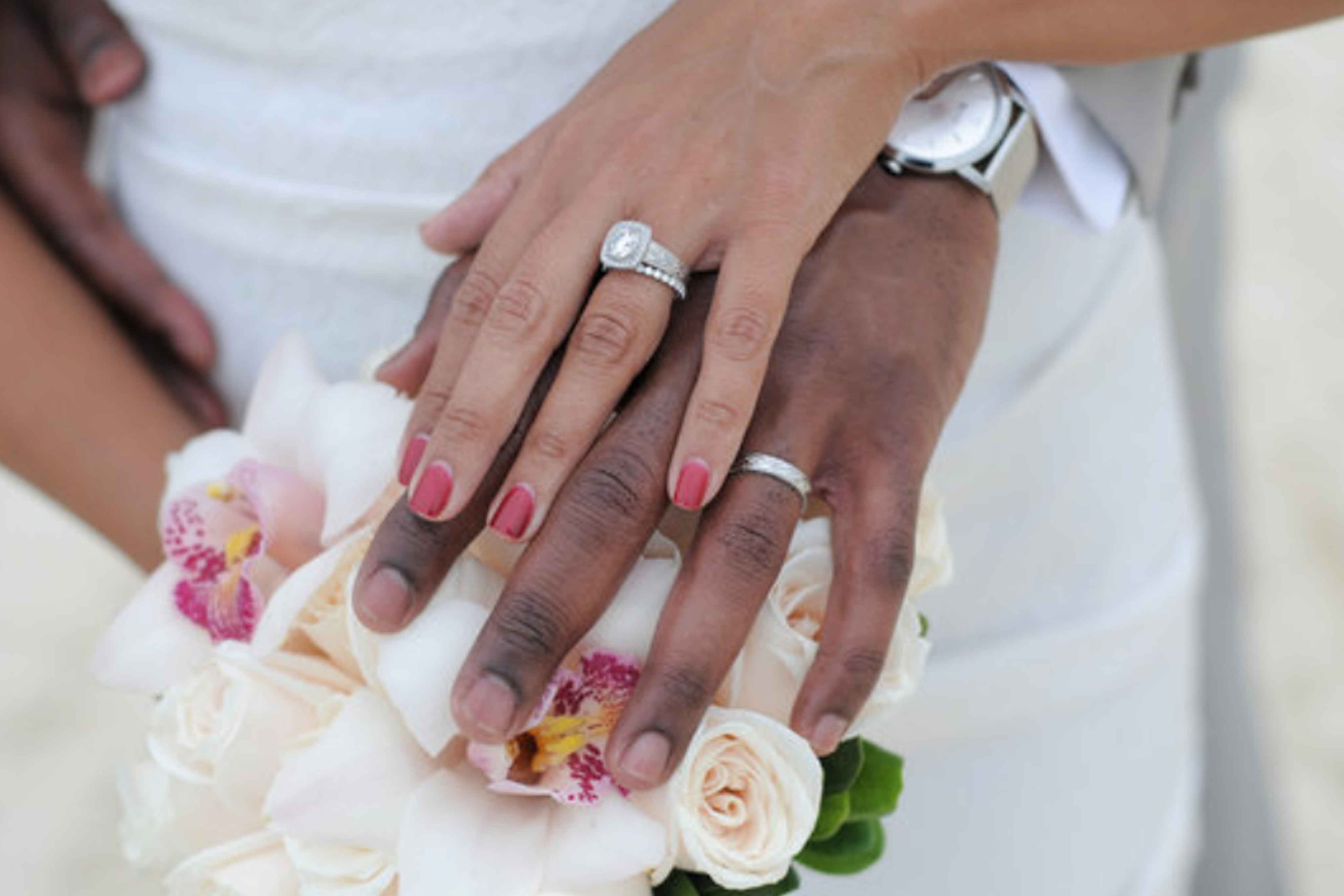 Close-up of a newlywed couple's hands with wedding rings, the bride holding a bouquet of white and pink flowers. The groom wears a white shirt and a watch, perfect for the homepage display.