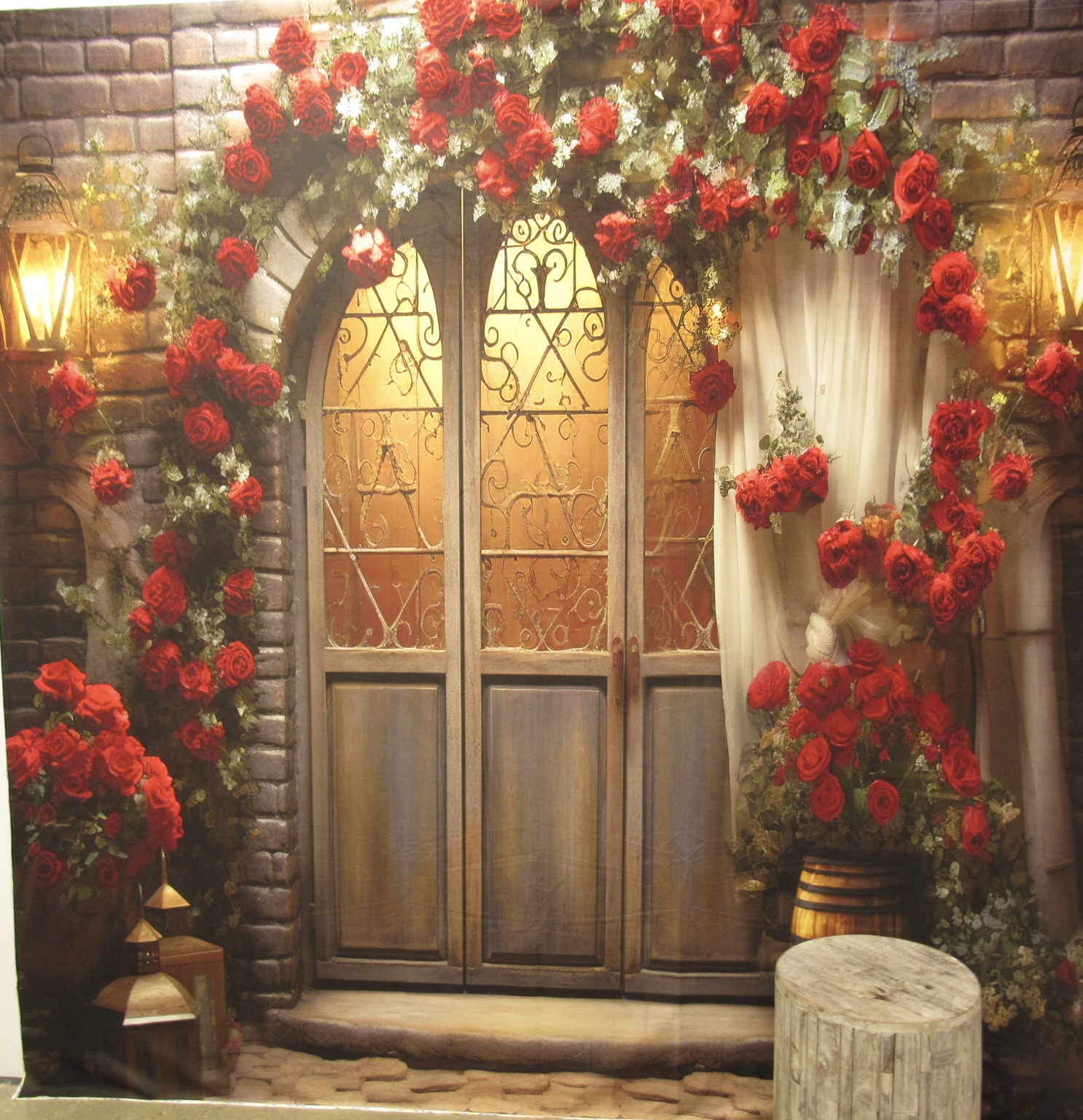 An ornate wooden double door with wrought-iron details flanked by lush red roses and greenery, set against a brick wall backdrop, accompanied by a white curtain and a wooden stool.