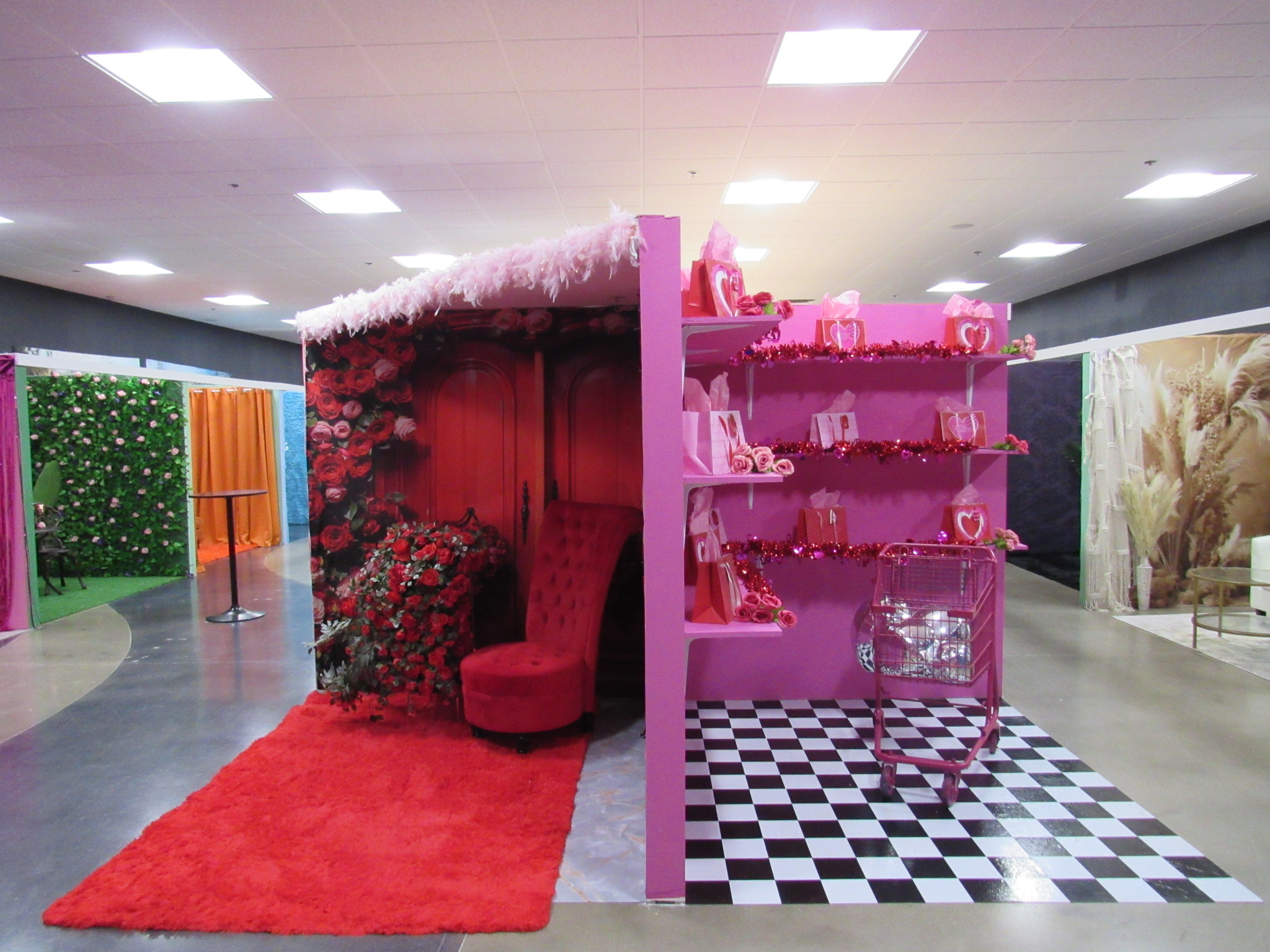A vibrant, whimsical display booth designed as a pink cabin with red and pink decorations, a red armchair, and shelves with pink items, set against a black and white checkered floor.