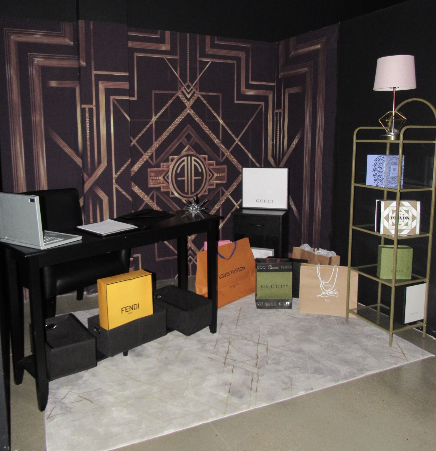 A luxury shopping-themed display with shopping bags from gucci, fendi, and other brands. a laptop sits on a black desk, and a lighted lamp stands beside a decorative shelf unit.