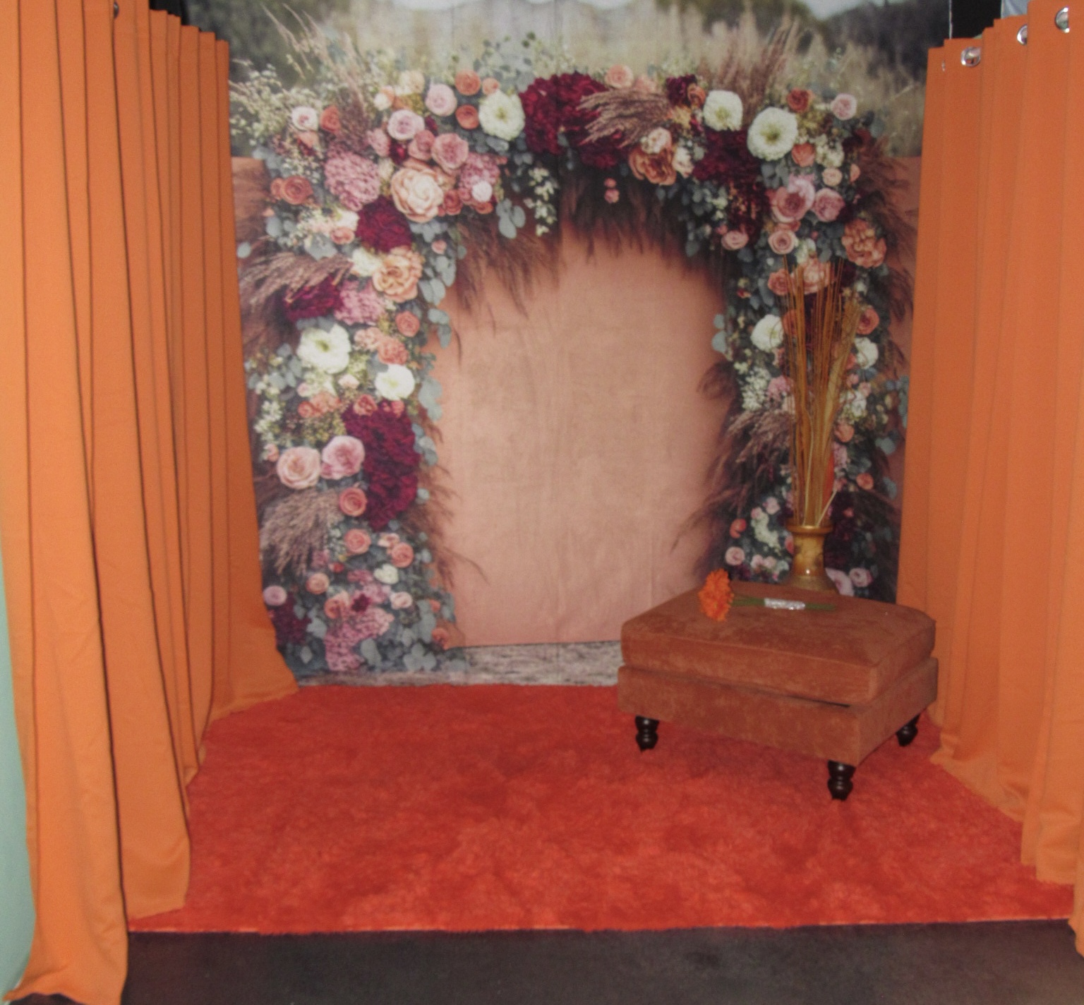 A cozy photo booth setup featuring orange curtains, a red carpet, and a floral arch with a variety of pink and white flowers. a small wooden stool sits in the center under the arch.