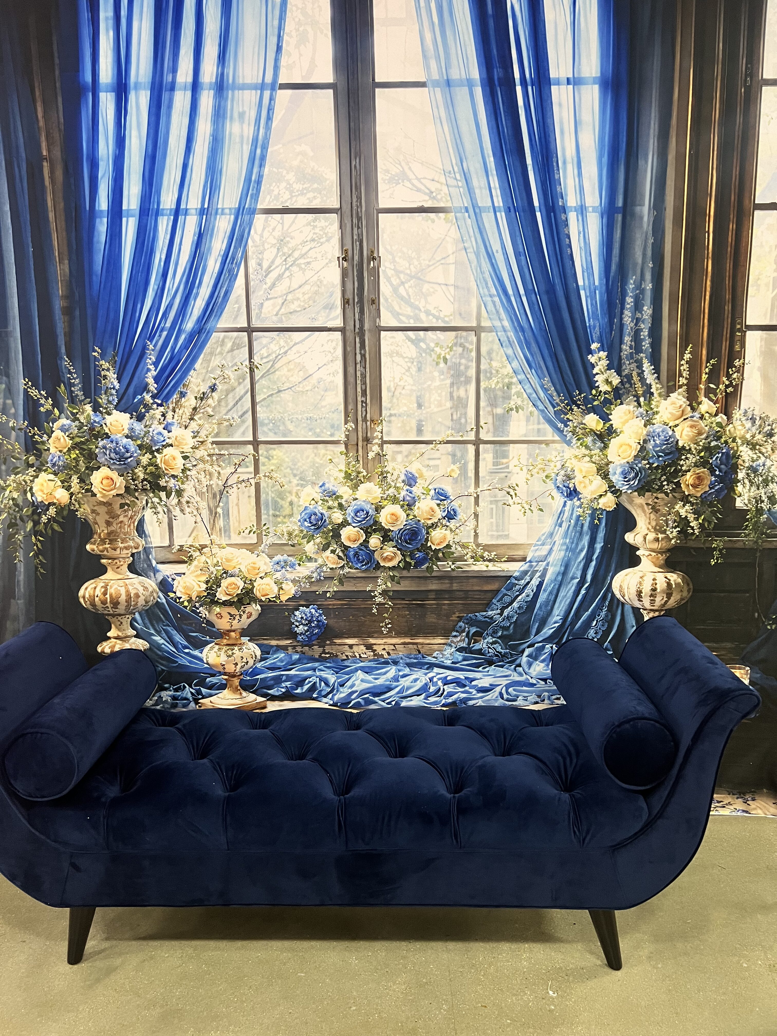 An elegant room with a large window draped with sheer blue curtains, a luxurious blue velvet sofa, and elaborate floral arrangements in gold vases on either side.