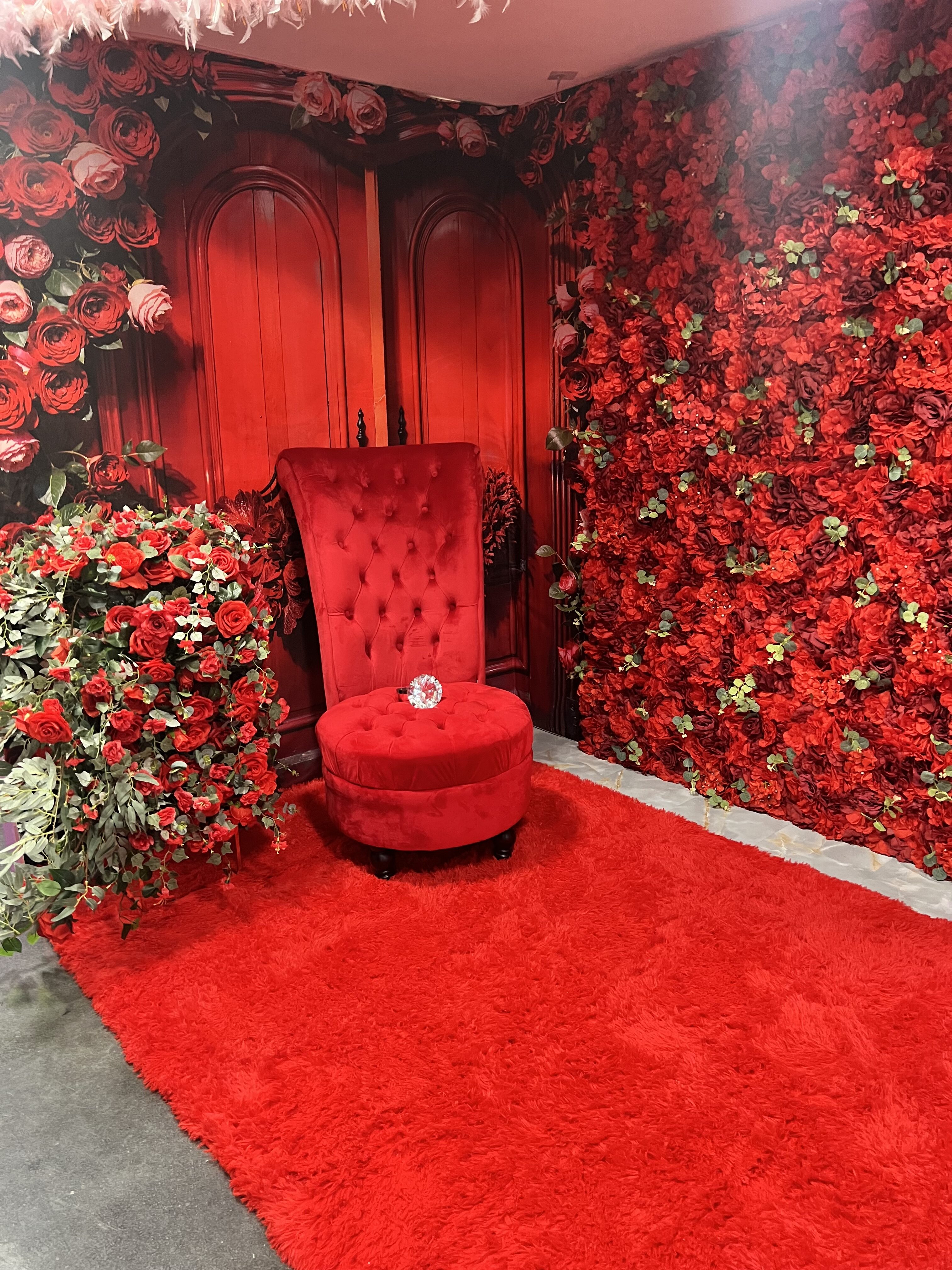 A lush red-themed room featuring walls adorned with red roses, a tufted red chair with a matching footstool, and a red carpet. red doors with brass handles are at the back.
