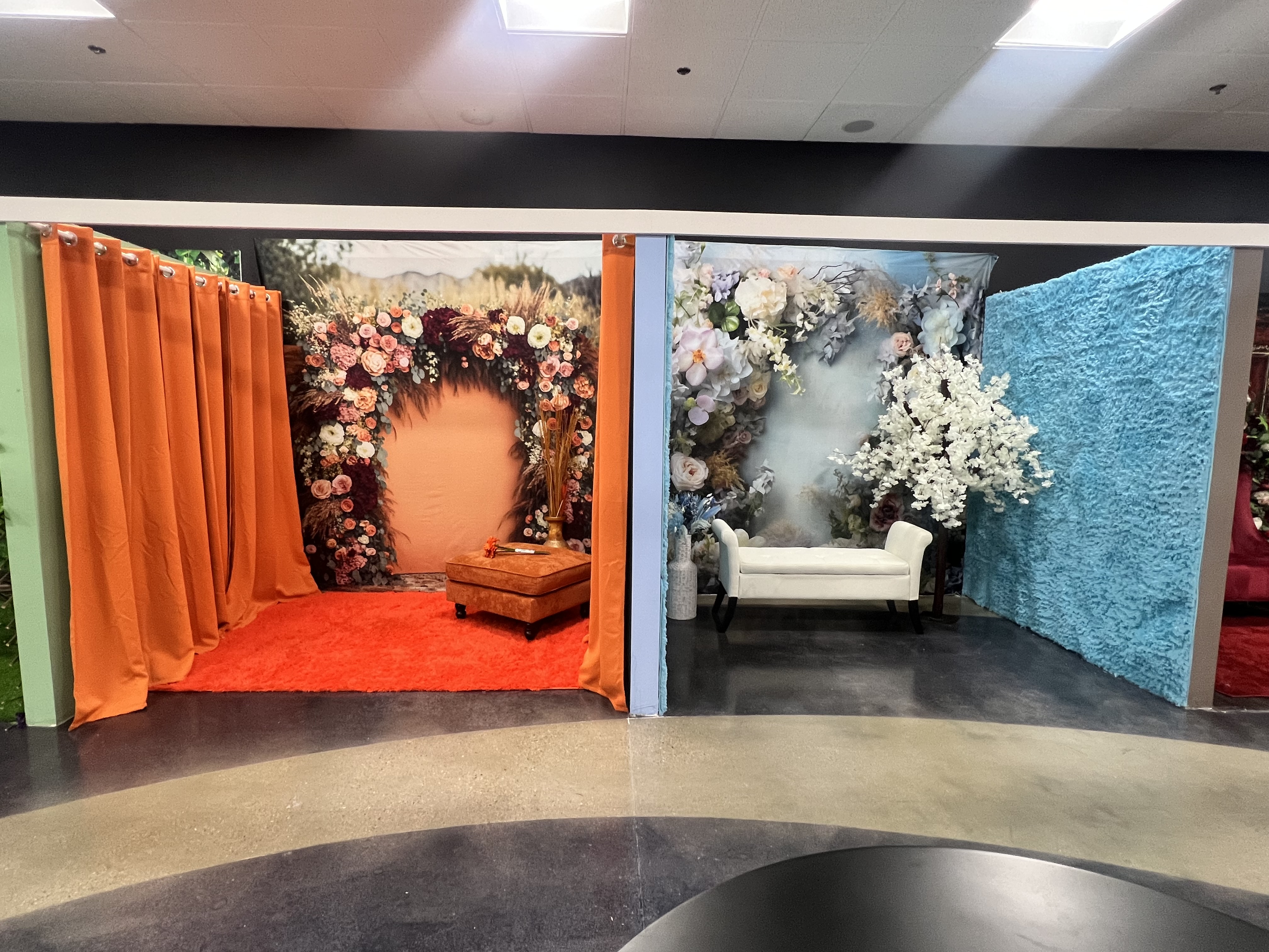 Two adjacent photo booth setups with floral backdrops: one with warm tones and orange curtains, the other with cool tones and baby blue curtains, each featuring seating options.