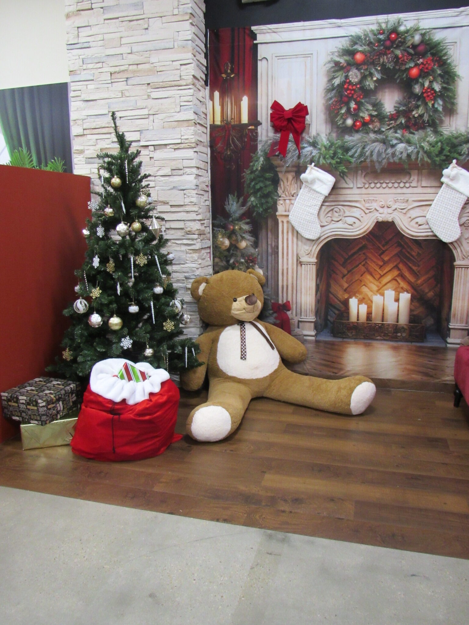 A decorated Christmas tree stands next to a large teddy bear, red gift bag, and wrapped present in front of a festive fireplace backdrop with stockings, candles, and holiday wreaths.