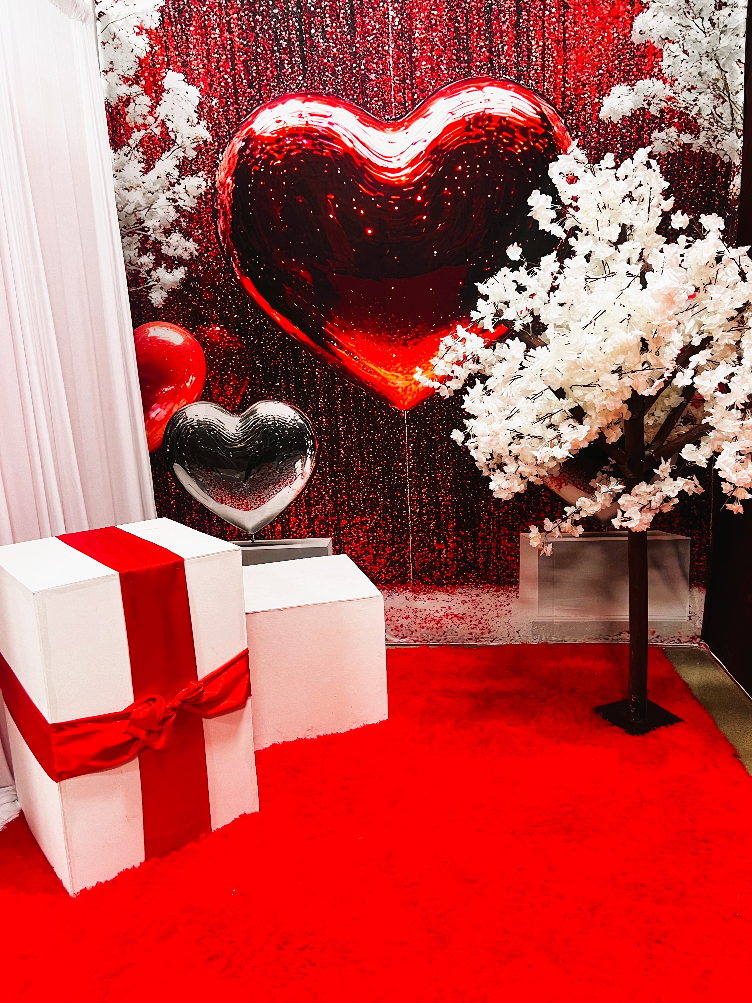 A festive scene with a white faux tree, large gift boxes wrapped in red ribbons, and a backdrop featuring oversized red and silver heart balloons with sparkling decorations. The floor is covered in bright red carpet.