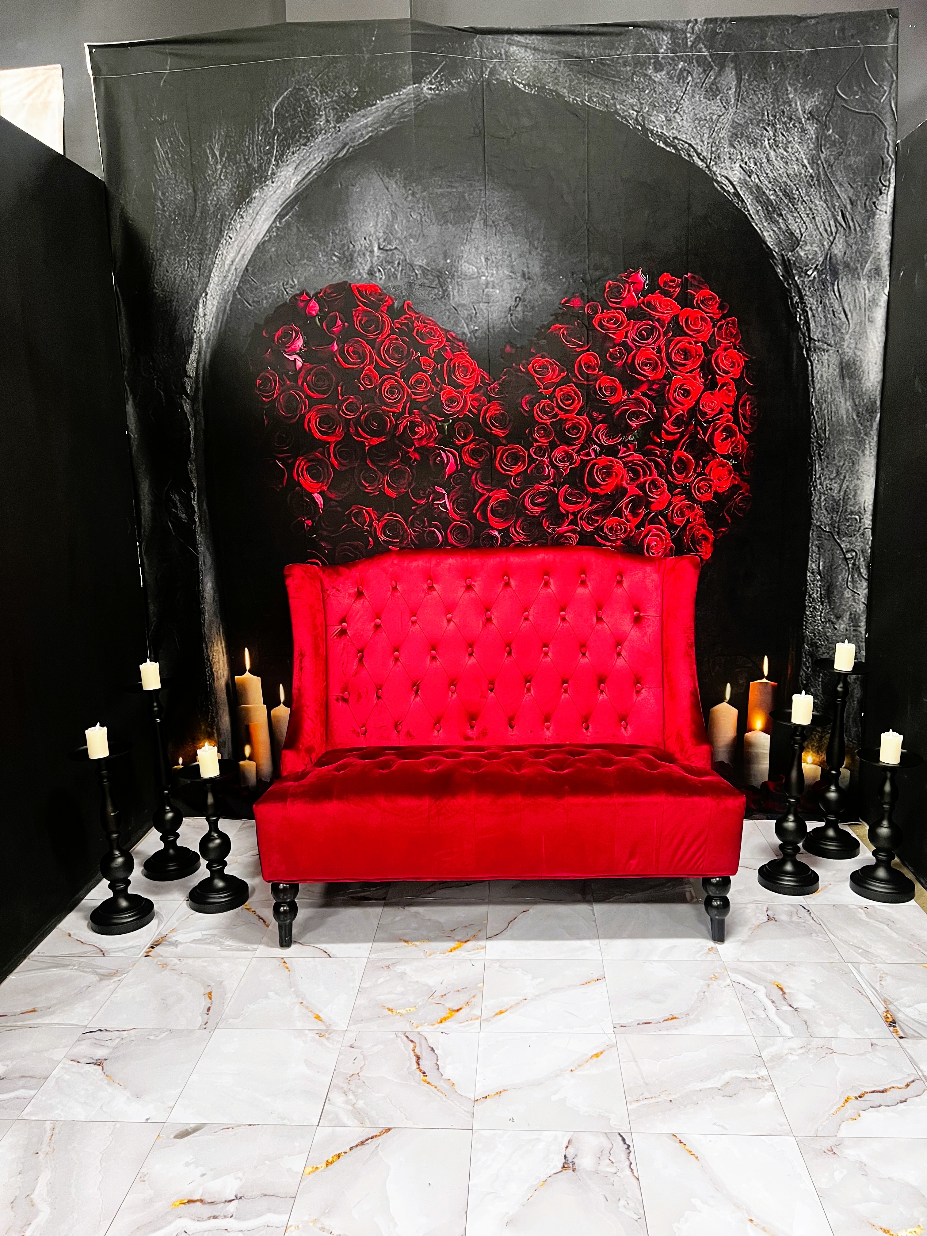 A red velvet tufted sofa sits in front of a black wall with a large red rose heart mural. Black candleholders with lit candles surround the sofa on a white marble floor.