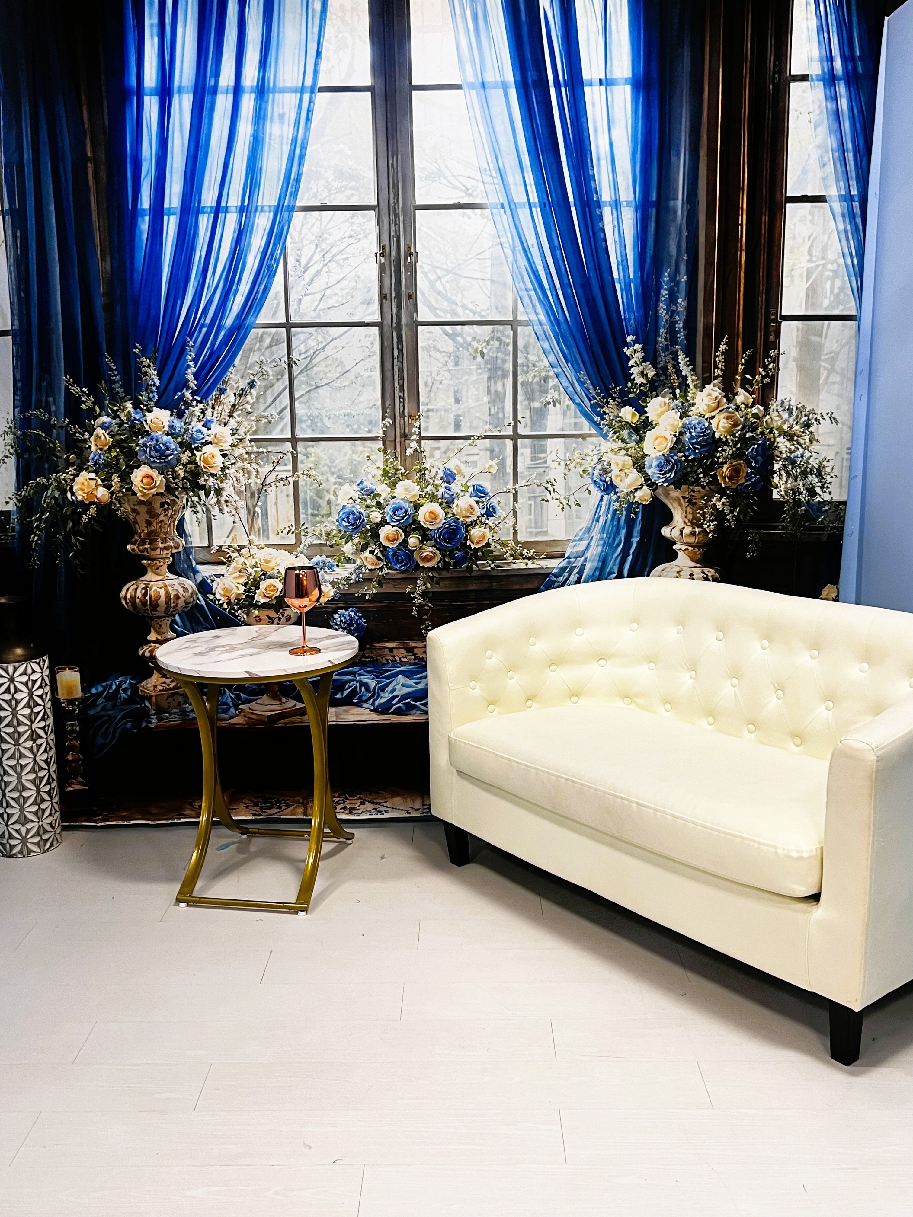 Elegant room with a white tufted loveseat, round marble side table with gold legs and a wine glass, large window with blue curtains, and floral arrangements with blue, white, and yellow flowers.