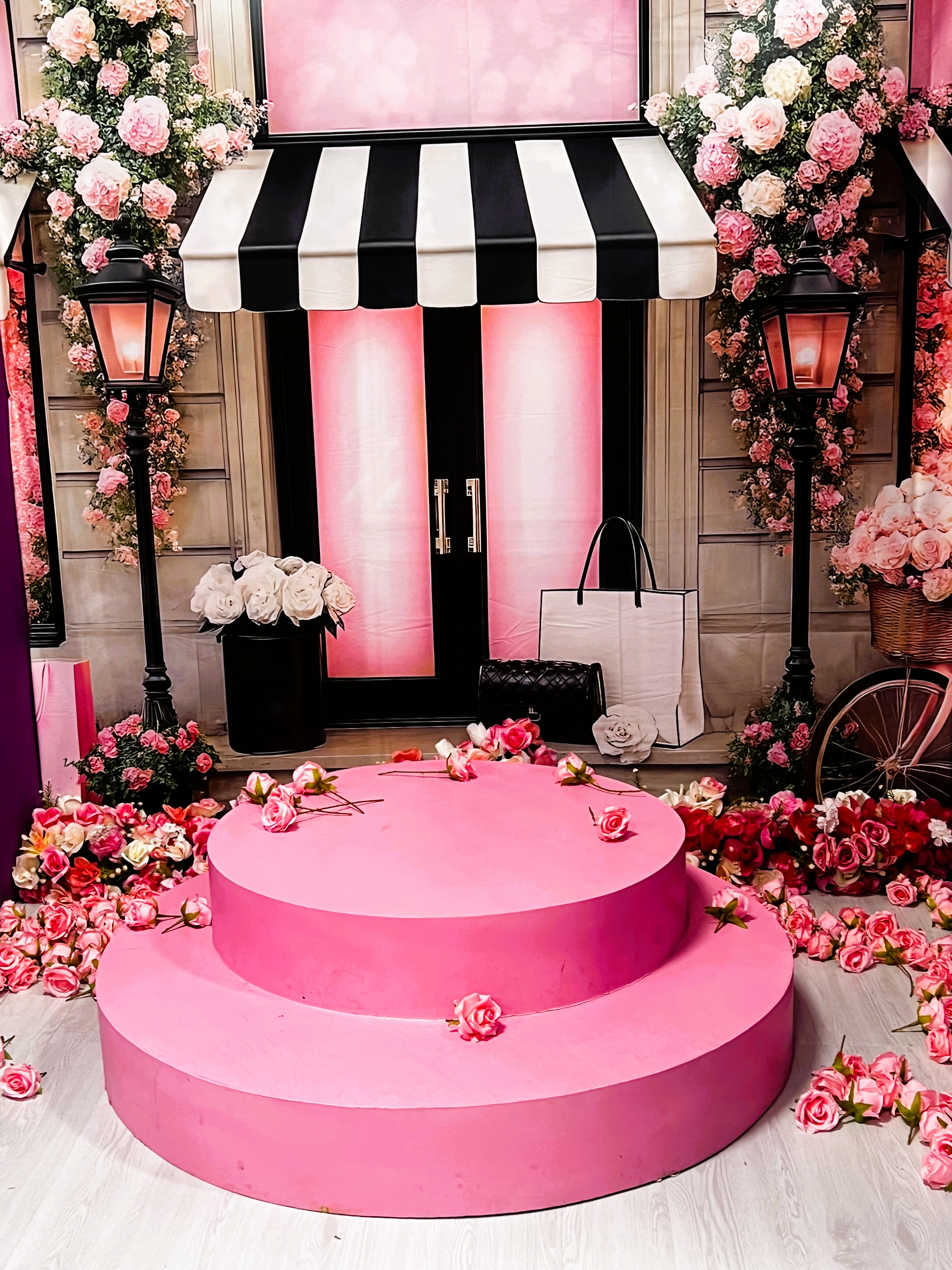 A pink tiered display platform is surrounded by scattered roses, floral arrangements, black lamps, a bicycle, and tote bags in front of a shop with black-and-white striped awning and pink accents.