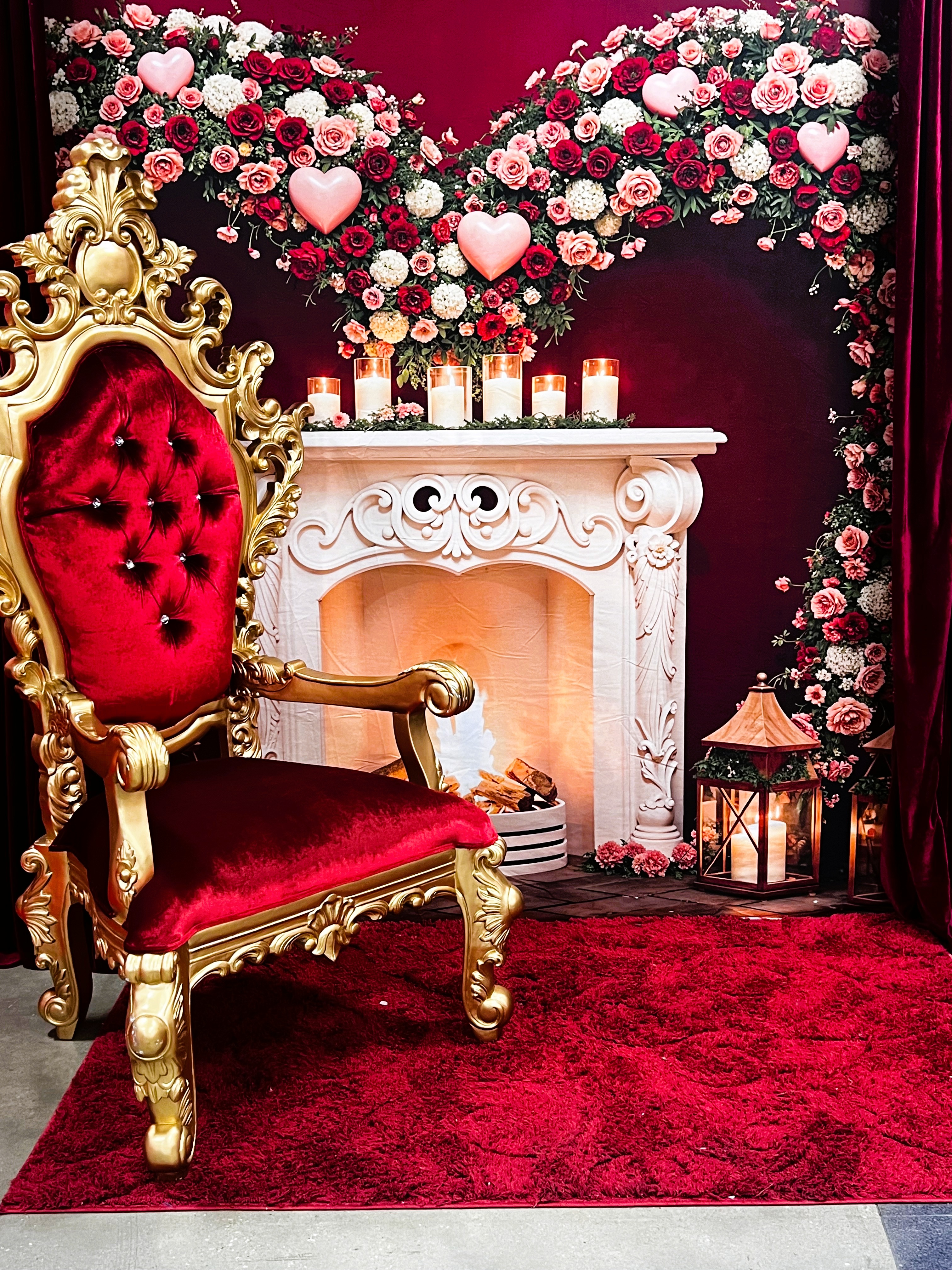 A luxurious gold and red velvet chair sits on a red rug in front of a white decorative fireplace, with lit candles and floral heart-shaped arrangements of red and pink roses in the background.