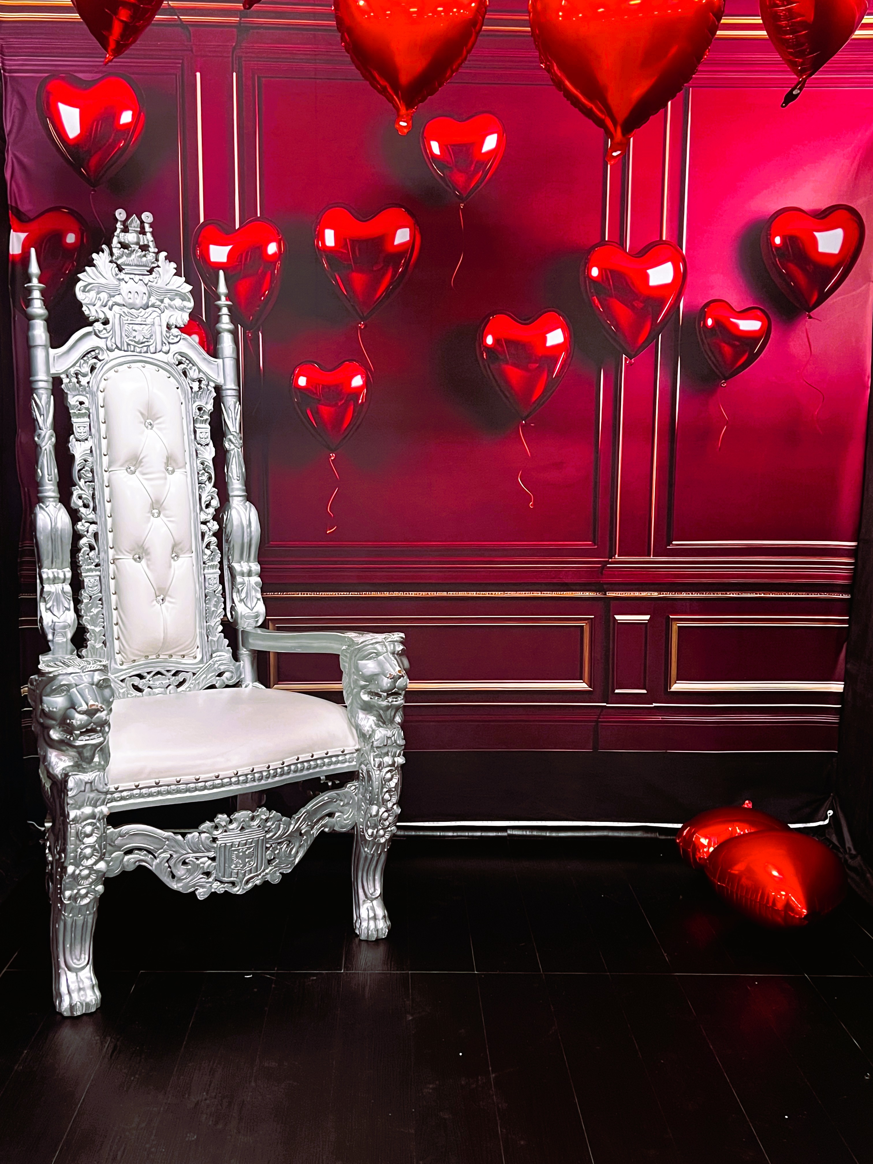 A tall, ornate silver throne sits beside a wall decorated with red, heart-shaped balloons against a rich red background. One balloon rests on the dark wooden floor.