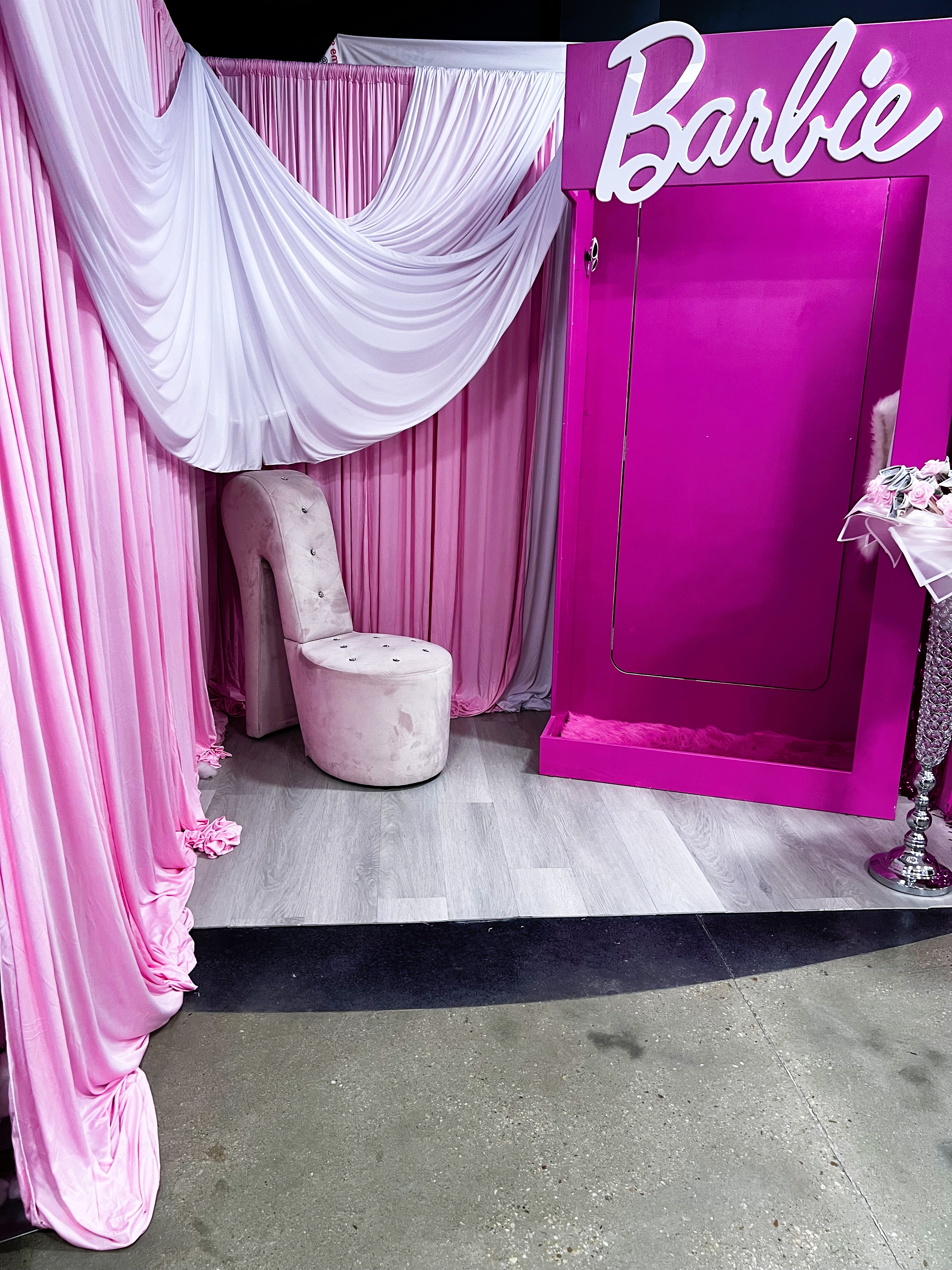 A Barbie-themed photo booth with pink and white draped curtains, a high-heeled shoe-shaped chair, a pink platform with “Barbie” written at the top, and a stand with pink flowers on the right.
