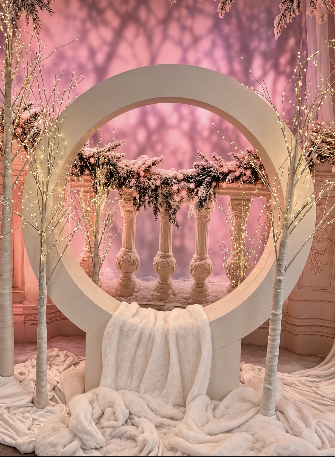A large white circular frame sits in front of a stone balustrade decorated with snowy evergreen branches, flanked by two lighted birch trees with a white faux fur blanket draped below.