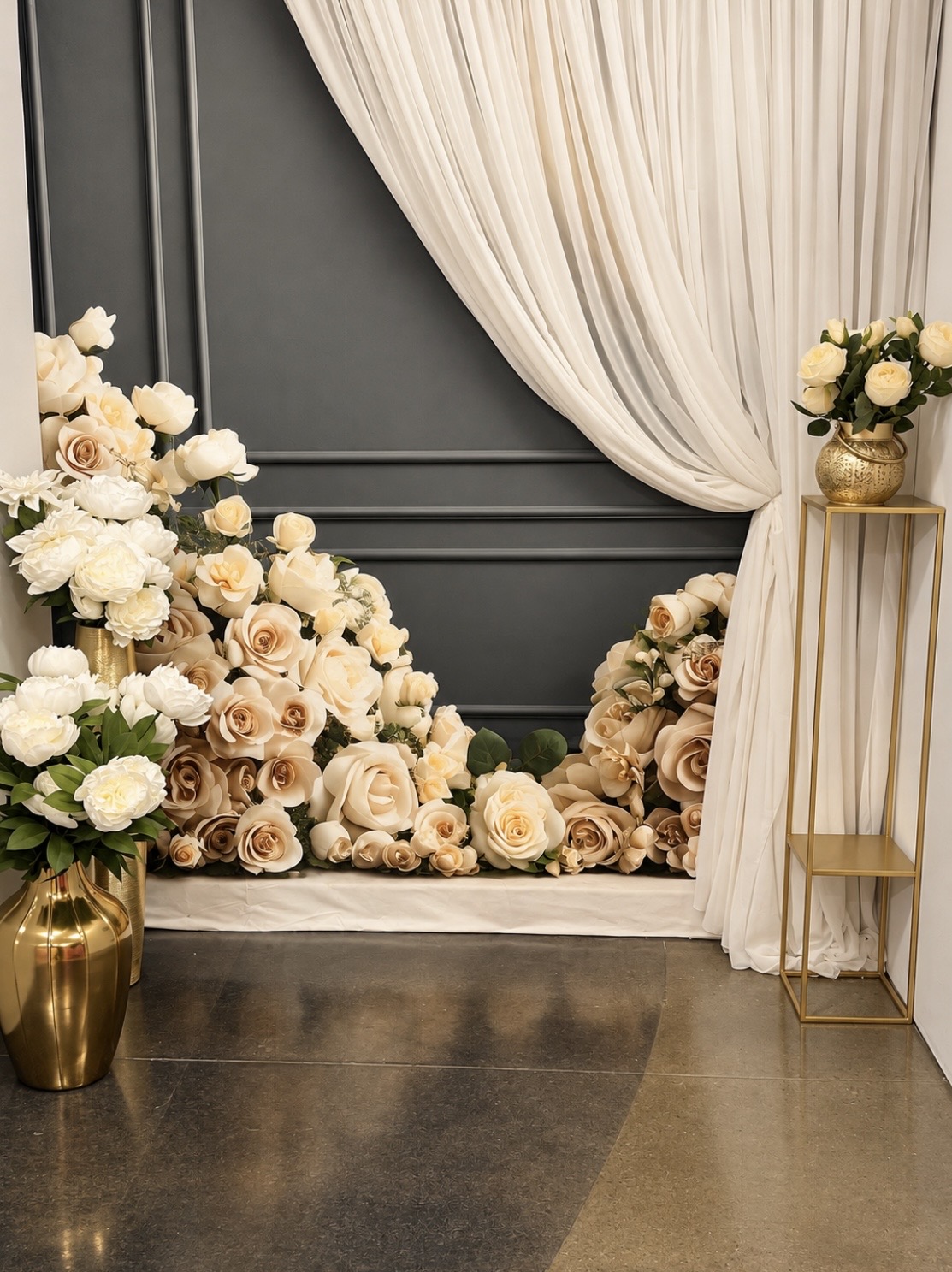 A charcoal-colored wall decorated with draped white curtains and a large arrangement of cream and white roses, with additional floral bouquets in gold vases on the floor and a gold stand.