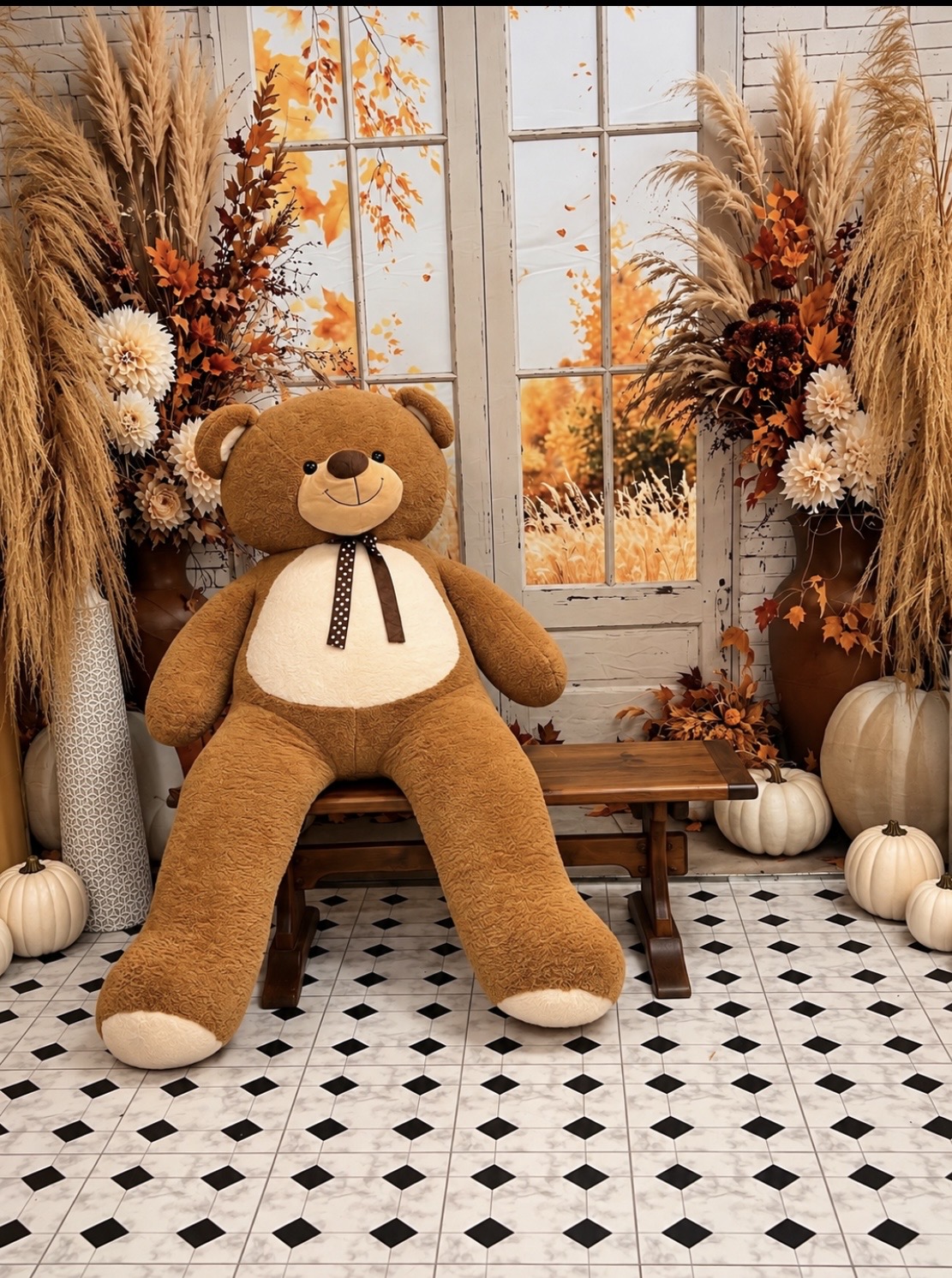 A large brown teddy bear with a brown ribbon sits on a wooden bench in front of a window decorated with autumn-themed flowers, dried grasses, and pumpkins.