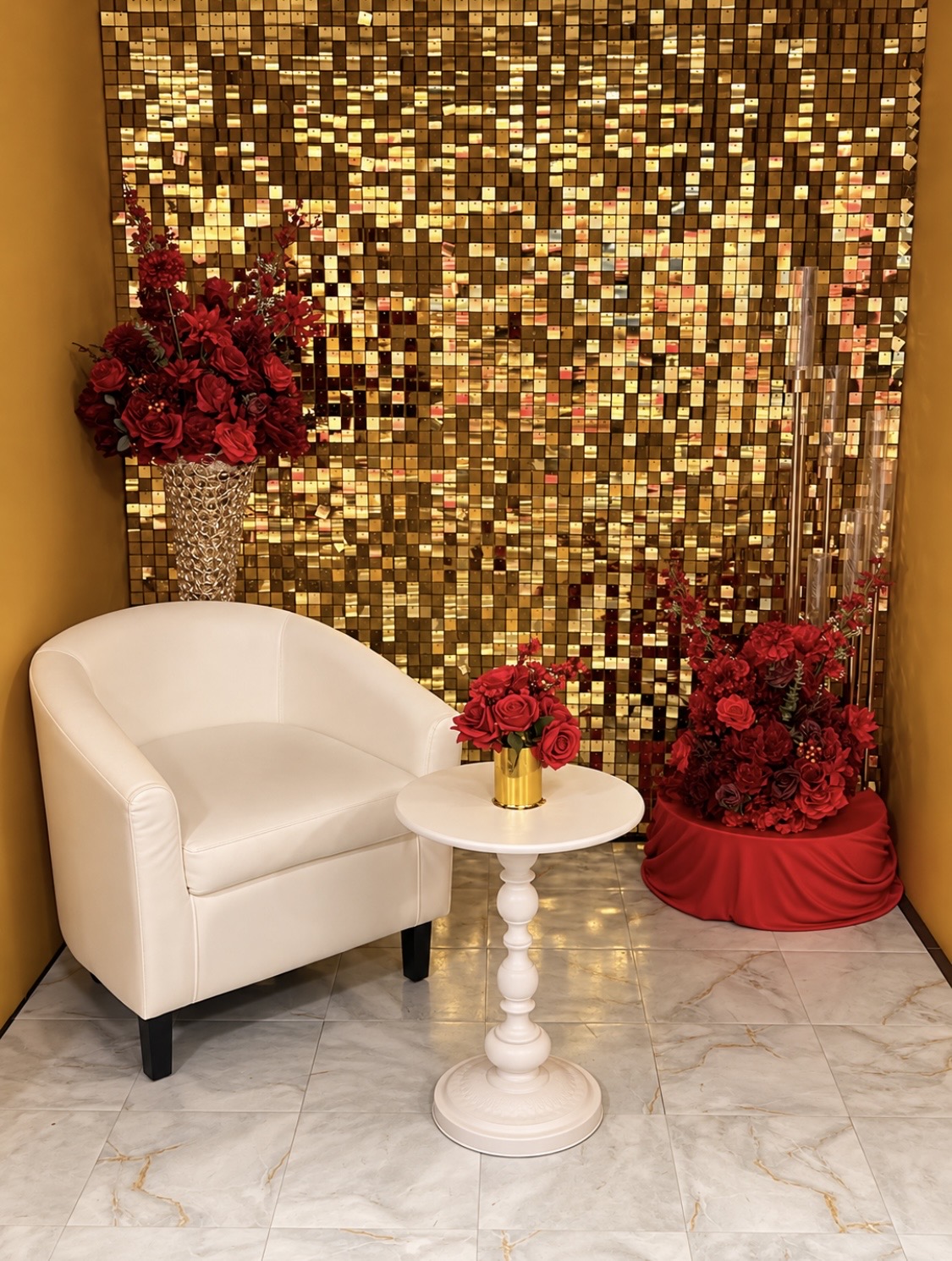 A white armchair and a small round table with a vase of red roses, set against a gold shimmery backdrop with additional floral arrangements.