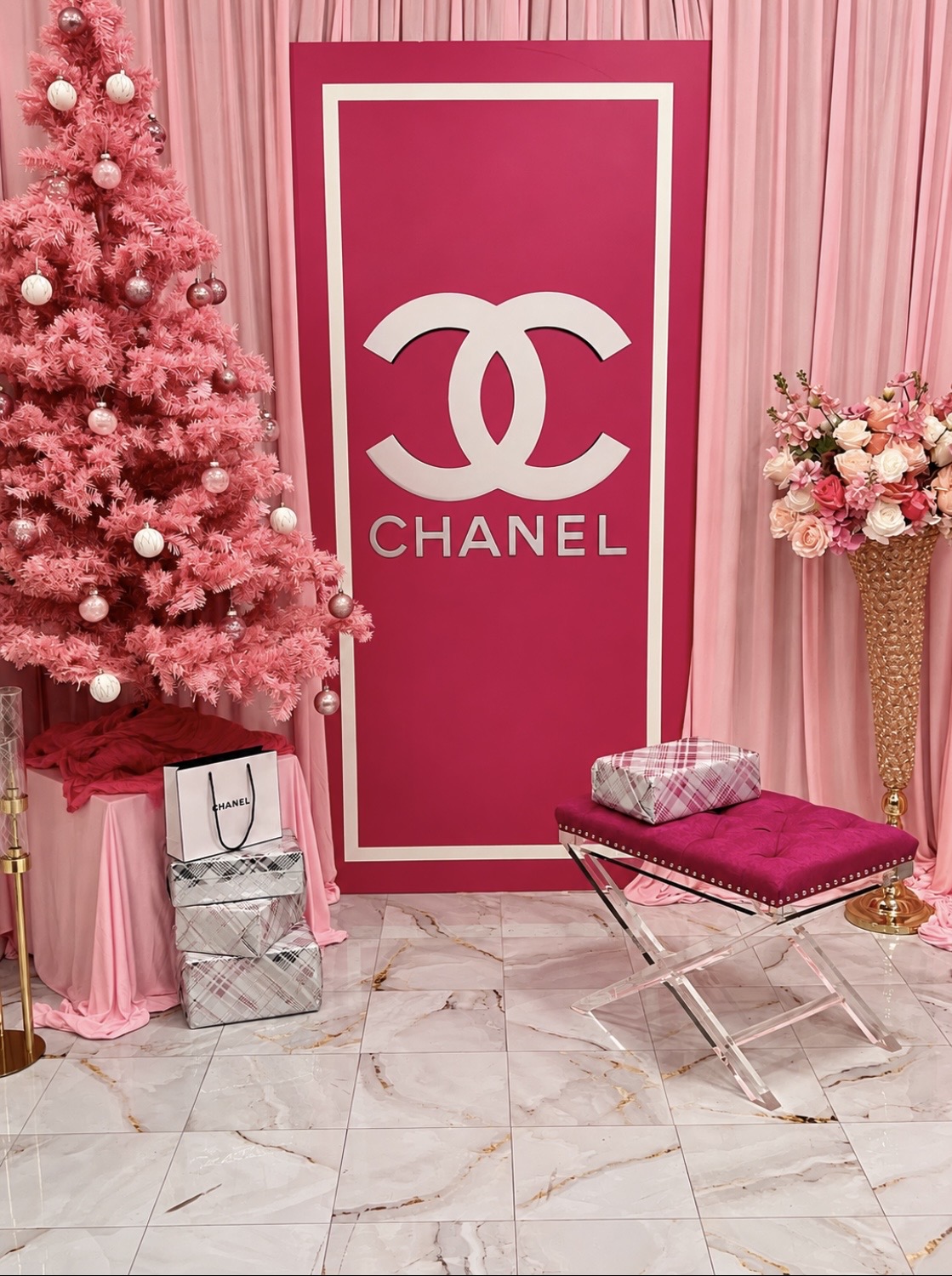 A pink Christmas tree and a Chanel-branded backdrop with wrapped gift boxes on the floor and a velvet bench.