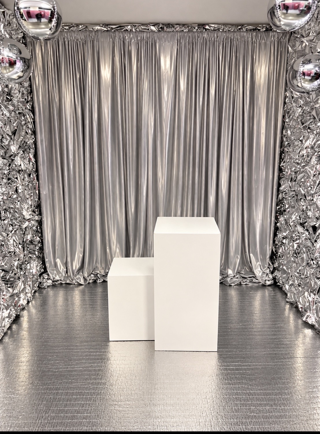 Two white rectangular display pedestals standing in front of silver metallic curtains and crinkled foil walls.